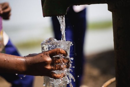 Photo for Closeup of a child hand collecting fresh drinking water from local tube-well. A hand of a kid collecting drinking water with a glass. Splash and sprinkle of water falling on glass. - Royalty Free Image
