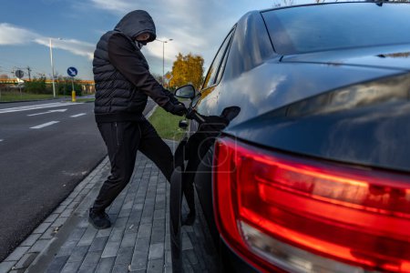 Foto de Intento de robar un coche usando una maleta en el centro de la ciudad - un ladrón en una capucha con una máscara en la cara - Imagen libre de derechos