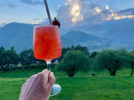 Photo for Hand holding red cocktail, Kazbegi and Gergeti Trinity Church at sunset in the background, Georgia. High quality photo - Royalty Free Image