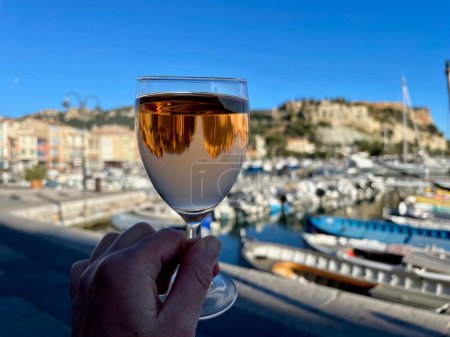 Photo for Female hand holding glass of rose wine, harbor and old town of Cassis in the background, Provence, France. High quality photo - Royalty Free Image