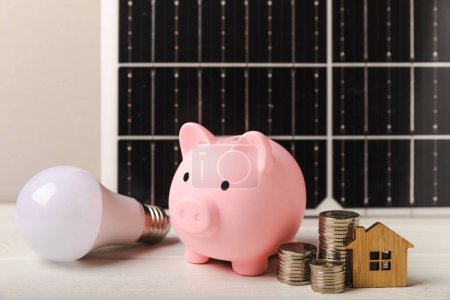 Geld, ein Sparschwein, eine LED-Lampe und ein Hausmodell auf dem Hintergrund von Sonnenkollektoren in Großaufnahme. Wirtschaftliche Vorteile erneuerbarer Energiequellen.