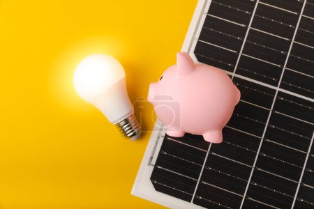 Composición plana con panel solar, lámpara led y alcancía sobre fondo amarillo. El concepto de ahorro de dinero y energía limpia. El concepto de ecología y desarrollo sostenible.
