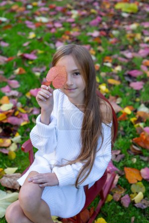 Photo for Autumn leisure, the need for vitamins in autumn, childrens entertainment. Portrait of a pretty girl with long hair covering half of her face with a red leaf on a blurred background - Royalty Free Image