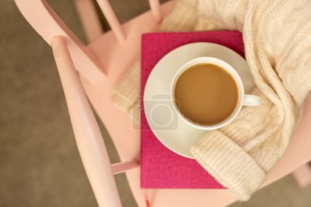 Photo for Hygge background in pink tones - a white mug with cocoa on a saucer, a dark pink book, a notebook, a warm sweater on a pink chair. The concept of rest, cozy home time - Royalty Free Image