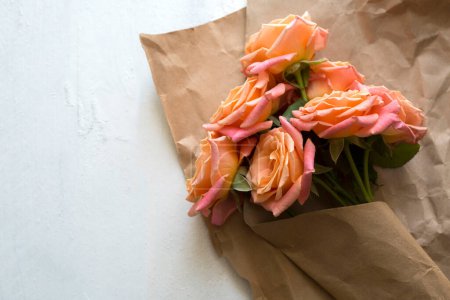 Foto de Regalo, flores, embalaje artesanal. Rosas de color rosa anaranjado en papel kraft sobre fondo blanco de madera - Imagen libre de derechos