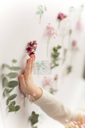 Photo for Exceptionality, choice, children. A childs hand touches a pink flower on a white wall with flowers. The concept of choice, exclusivity. Floral decoration - Royalty Free Image