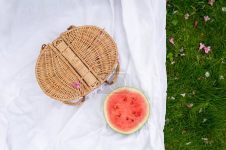 Téléchargez les photos : Pique-nique, goûter, été. Panier de pique-nique et pastèque sur une couverture blanche avec une bande verte d'herbe et de fleurs - en image libre de droit