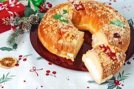 Roscon de reyes mit Sahne und Weihnachtsschmuck auf einem roten Teller über der Weihnachtstischdecke. Kings Day Konzept spanischer Dreikönigskuchen. Typisches spanisches Dessert zu Weihnachten