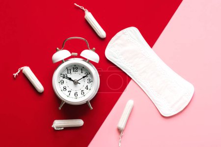 Top view of sanitary pad, menstrual tampons and white alarm clock over pink and red background. Concept of menstruation and menopause