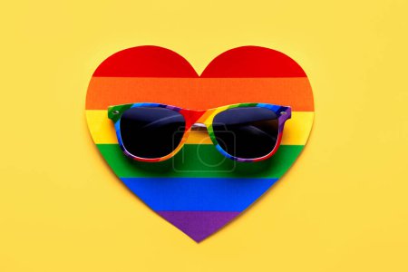 Closeup of LGTBI flag sunglasses on LGTBI heart over yellow background. LGBT Pride concept