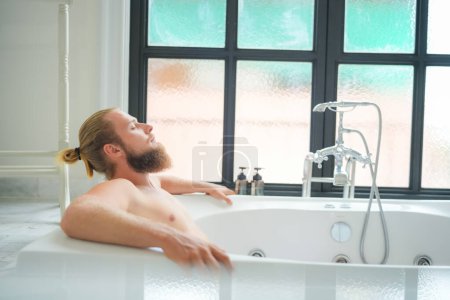 Foto de Hombre joven relajado bañándose en el moderno baño interior de lujo. Spa, bienestar, concepto de cuidado corporal - Imagen libre de derechos