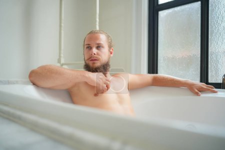 Téléchargez les photos : Jeune homme détendu se baignant dans la salle de bain moderne intérieur de luxe. Spa, bien-être, soins du corps concept - en image libre de droit