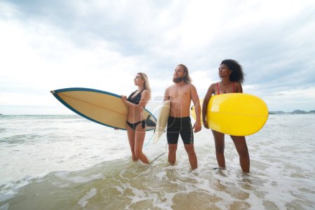 Photo for Young mixed race women and a Caucasian women and man enjoying their time at the beach with their friends, holding surfboards - Royalty Free Image