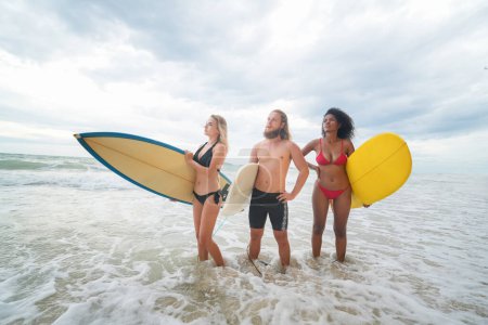 Téléchargez les photos : Jeunes femmes métisses et une femme et un homme caucasiens profitant de leur temps à la plage avec leurs amis, tenant des planches de surf - en image libre de droit