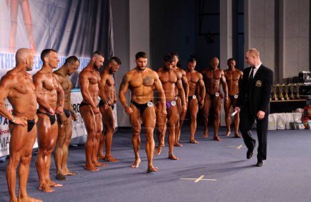 Photo for Man assistant judge helping group of muscly men posing on podium, bodybuilding. Kyiv championship among amateurs. December 5, 2019. Kyiv, Ukraine - Royalty Free Image