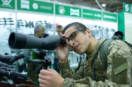 Photo for Young man in military uniform looking through portable monocular telescope, gun shop. October 08, 2019. Kiev, Ukraine - Royalty Free Image