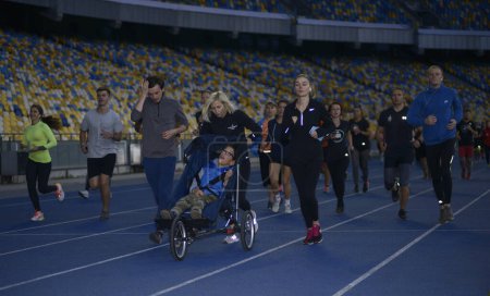 Photo for Group of people running on tracks of the stadium, in the center - woman running and pushing the wheelchair with boy with special needs (cerebral palsy). October 14, 2019. Kiev, Ukraine - Royalty Free Image