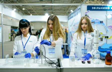 Foto de Tres mujeres jóvenes asistentes de laboratorio que trabajan con ionómetro y valorador para medir la calidad del agua potable. Exposición Industrial Ucrania. 6 de noviembre de 2019. Kiev, Ucrania - Imagen libre de derechos