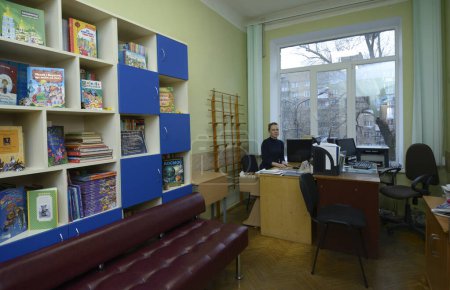 Foto de Bibliotecaria sentada en la mesa de trabajo de la biblioteca para alumnos de primaria, libros infantiles en estanterías. 3 de marzo de 2020. Kiev, Ucrania - Imagen libre de derechos