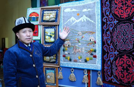 Téléchargez les photos : Homme asiatique dans un chapeau kirghize indigène pointant sur une broderie traditionnelle kirghize colorée suzani, tapis. Festival de la culture orientale. 20 mars 2018. Kiev, Ukraine - en image libre de droit