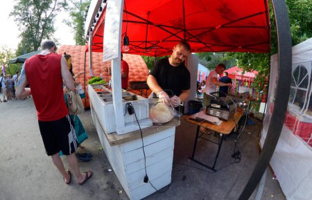 Foto de El hombre cocina comida en el puesto de comida de la calle, la gente de pie en una cola, campamento de festejos. Festival Vedalife. 7 de agosto de 2017. Kiev, Ucrania - Imagen libre de derechos