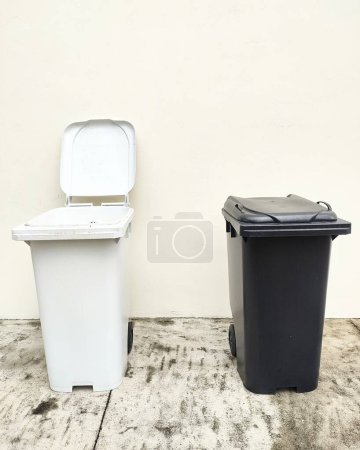 One white and one black garbage bins side by side in front of a white wall. Example of urban waste management and recycling.
