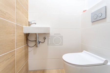 Photo for Modern bathroom with a minimalist design, featuring a small white wall-mounted sink with a silver faucet on beige wood-effect tiled walls, white toilet with a flush button panel on the wall beside it. - Royalty Free Image