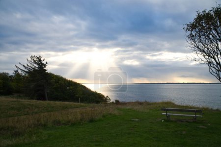 Photo for On the coast of Hundested. Sun rays break through the dramatic sky through the clouds. Meadow with park bench in foreground. Landscape shot in Denmark - Royalty Free Image