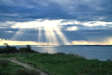 Photo for On the coast of Hundested. Sun rays break through the dramatic sky through the clouds. Meadow with path in foreground. Landscape shot in Denmark - Royalty Free Image