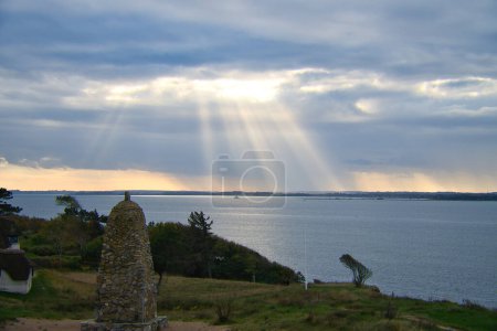 Photo for On the coast of Hundested. Sun rays break through the dramatic sky through the clouds. Meadow with stone tower in the foreground. Landscape shot in Denmark - Royalty Free Image