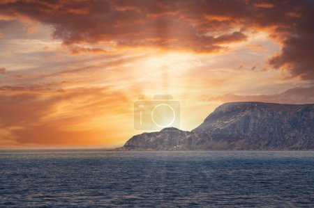 View from the sea to the West Cape in Norway at sunset. Sun shines over the rocks. Waves in the foreground, rocks in the background. Westernmost point of Europe mainland.