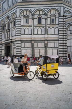 Foto de Florencia, Toscana / Italia - 15 de junio de 2019: Dos conductores de rickshaw sentados en su velo-taxi frente al Baptisterio de San Juan (Battistero di San Giovanni) en Florencia esperando a los turistas y hablando entre sí - Imagen libre de derechos