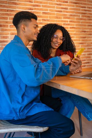 Téléchargez les photos : Un couple multiethnique utilisant un téléphone portable se fait sourire à la maison, un petit ami latino et une petite amie afro-américaine sur les médias sociaux essayer une nouvelle application mobile. Les jeunes travaillent avec un ordinateur portable - en image libre de droit