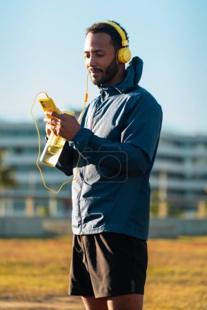 Téléchargez les photos : Homme athlétique avec casque fatigué de l'exercice prend une pause pour boire de l'eau tout en utilisant une application de téléphone portable à l'extérieur. - en image libre de droit