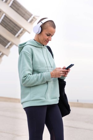 Téléchargez les photos : Une femme sportive avec casque fatiguée de l'exercice prend une pause pour utiliser une application de messagerie de téléphone portable. - en image libre de droit