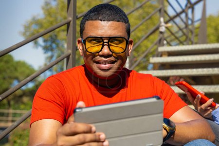 Happy latin man wearing sunglasses smiling while surfing the internet with digital tablet looking at camera outdoors.