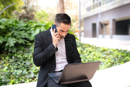 Middle Aged Businessman Dressed in Formal Attire Having A Call With Phone, Working With Laptop