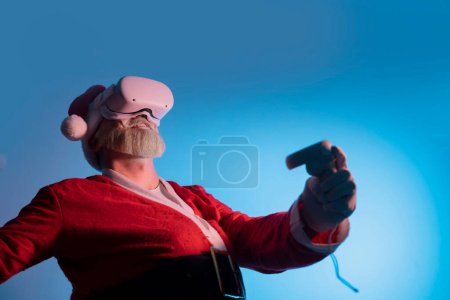 Téléchargez les photos : Santa porter vr casque gadget appareil équipement regarder l'expérience réaliste. Les personnes âgées moustache aux cheveux gris barbu Père Noël l'expérience de la réalité virtuelle jouer dans le jeu vidéo - en image libre de droit