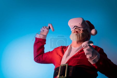 Téléchargez les photos : Santa porter vr casque gadget appareil équipement regarder l'expérience réaliste. Les personnes âgées moustache aux cheveux gris barbu Père Noël l'expérience de la réalité virtuelle jouer dans le jeu vidéo - en image libre de droit