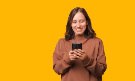 Téléchargez les photos : Photo de femme heureuse en sweat à capuche utilisant un smartphone sur fond jaune. - en image libre de droit