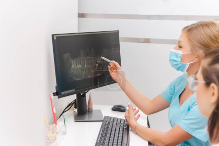 Mujer dentista con máscara de protección explica a la paciente la toma de rayos X de la pantalla de la computadora.