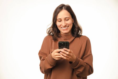 Foto de Mujer extática está usando o escribiendo en su teléfono mientras sonríe ampliamente en un estudio blanco. - Imagen libre de derechos