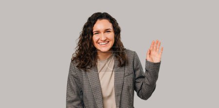 Foto de Photo of charming happy young woman in casual suit making Hello gesture over grey background. - Imagen libre de derechos