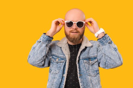 Téléchargez les photos : Handsome bearded bald man wearing jeans is holding cool white sunglasses over yellow backdrop. - en image libre de droit
