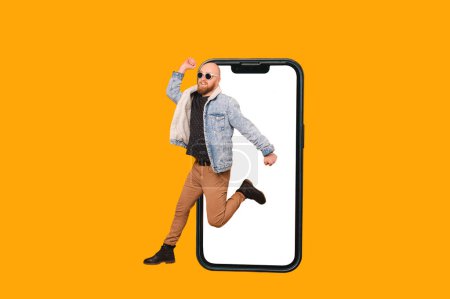 Photo for Young bald smiling bearded man wearing sunglasses is jumping out of a phone over yellow background. - Royalty Free Image