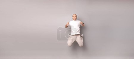 Photo for Banner size studio shot of a young man wearing headphones and jumping high over grey background. - Royalty Free Image