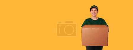 Photo for Banner size photo with copy space of a delivery woman holding a big box over yellow background. - Royalty Free Image