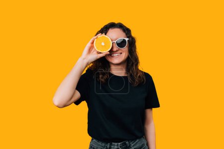Téléchargez les photos : Une femme souriante portant des lunettes couvre un œil d'une demi-orange sur fond jaune. - en image libre de droit