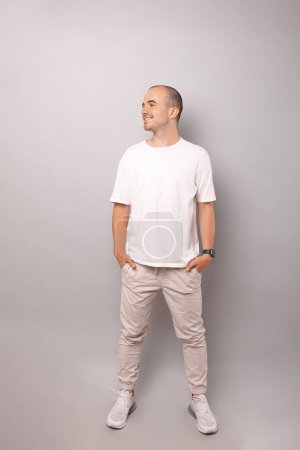 Photo for Full body shot of a bald looking aside man standing with hands in pockets over grey background. - Royalty Free Image