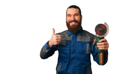 Photo for Handsome smiling handy man is holding an angle grinder and a thumb up. Studio shot over white background. - Royalty Free Image